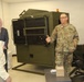 A U.S. Army soldier in MultiCam/OCP camouflage uniform is briefing or presenting to two civilians in an indoor facility. The soldier appears to be explaining a large olive-drab military equipment unit, possibly a ruggedized electronics or communications shelter. The setting appears to be a demonstration or tour of military equipment.