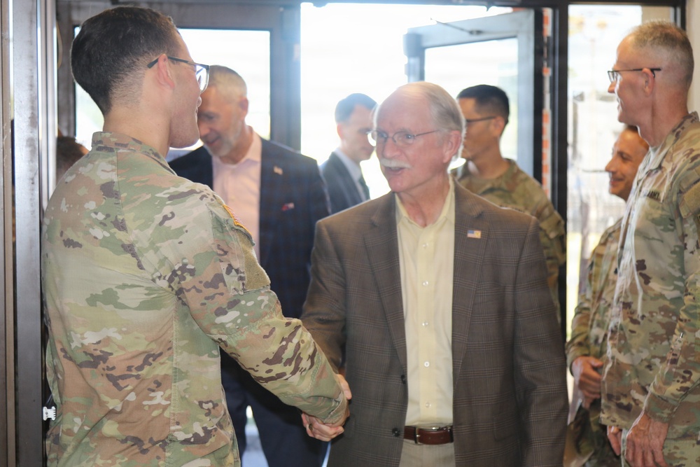 A civilian man in a brown blazer wearing an American flag lapel pin is shaking hands with a U.S. Army soldier in OCP (Operational Camouflage Pattern) uniform. The setting appears to be a military installation or facility, with several other soldiers and civilians visible in the background. The interaction suggests an official visit or greeting ceremony.