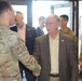 A civilian man in a brown blazer wearing an American flag lapel pin is shaking hands with a U.S. Army soldier in OCP (Operational Camouflage Pattern) uniform. The setting appears to be a military installation or facility, with several other soldiers and civilians visible in the background. The interaction suggests an official visit or greeting ceremony.
