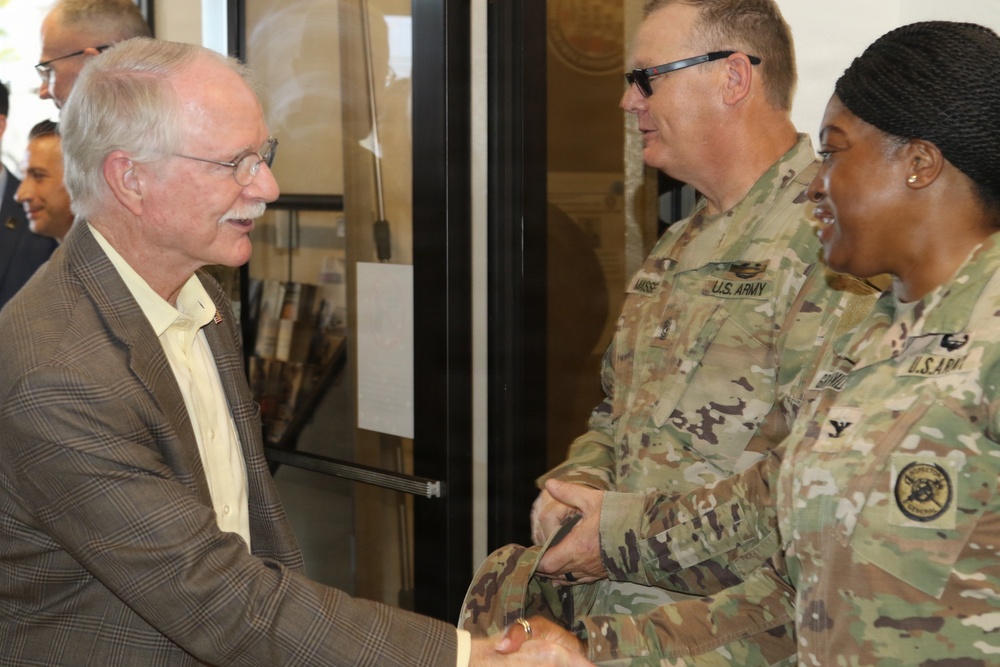 A civilian man in a plaid blazer is shaking hands with a U.S. Army female soldier, while a male U.S. Army soldier stands nearby smiling. The setting appears to be an indoor facility entrance or lobby. The interaction suggests a formal greeting or official visit.