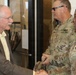 A civilian man in a plaid blazer is shaking hands with a U.S. Army female soldier, while a male U.S. Army soldier stands nearby smiling. The setting appears to be an indoor facility entrance or lobby. The interaction suggests a formal greeting or official visit.