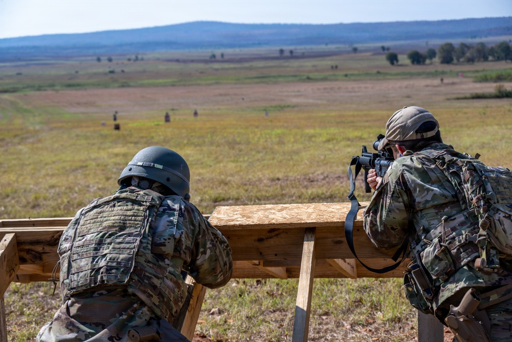 Two military personnel in camouflage uniforms are positioned at a wooden shooting bench on an outdoor firing range. One soldier is actively aiming a rifle equipped with an optic scope downrange, while the other observes. Multiple targets are visible across the open field in the background.