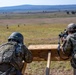 Two military personnel in camouflage uniforms are positioned at a wooden shooting bench on an outdoor firing range. One soldier is actively aiming a rifle equipped with an optic scope downrange, while the other observes. Multiple targets are visible across the open field in the background.