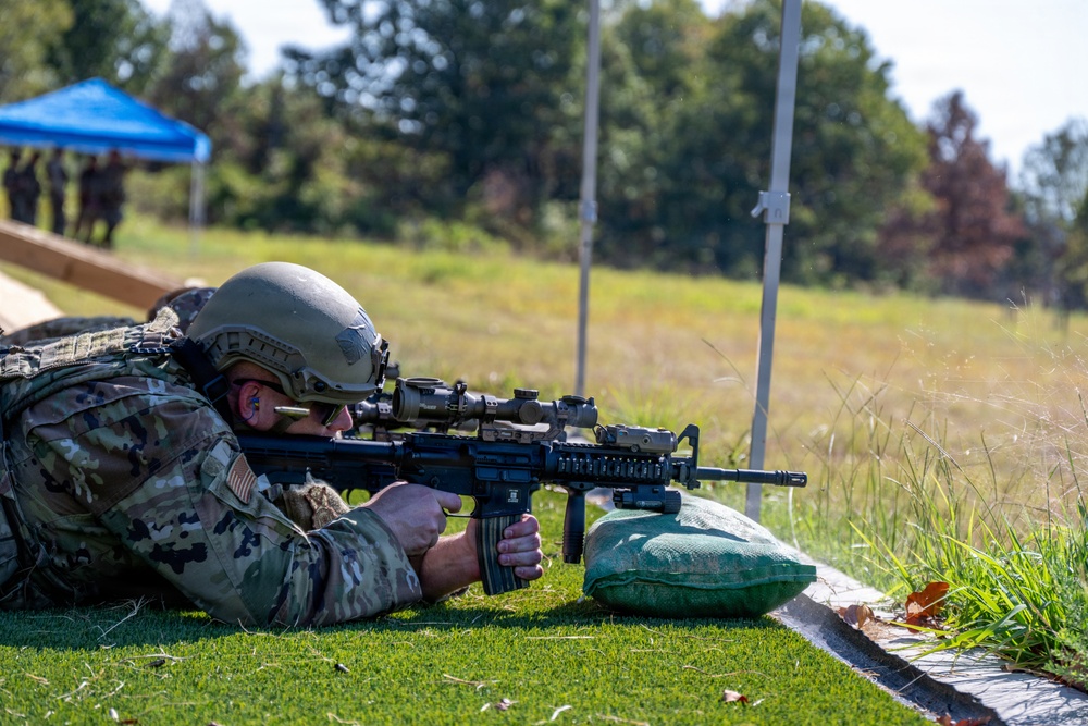 A soldier in full combat gear is in a prone firing position on a grass range, aiming a rifle equipped with optics and accessories. The setting appears to be a military marksmanship training or qualification range, with other personnel visible in the background near a blue canopy. The image captures a controlled shooting exercise in an outdoor environment.