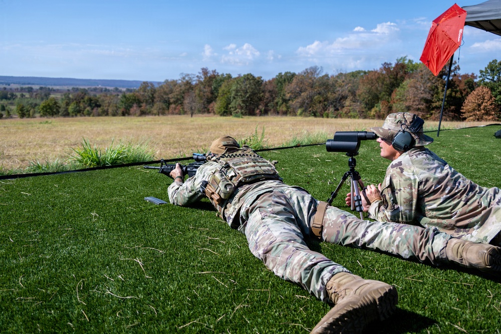Two military personnel in multicam camouflage uniforms are positioned prone on a grass shooting range. One soldier is firing a long-range rifle while the other acts as a spotter using a spotting scope mounted on a tripod. The setting appears to be an outdoor range during autumn, with trees showing fall foliage in the background.