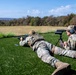 Two military personnel in multicam camouflage uniforms are positioned prone on a grass shooting range. One soldier is firing a long-range rifle while the other acts as a spotter using a spotting scope mounted on a tripod. The setting appears to be an outdoor range during autumn, with trees showing fall foliage in the background.