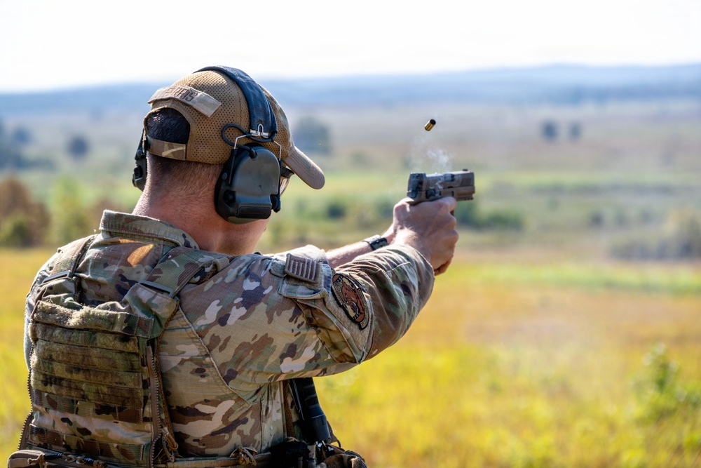 A military or law enforcement personnel is firing a semi-automatic pistol at an outdoor shooting range. A spent brass casing is visibly ejecting from the firearm, captured mid-air by the camera. The individual is wearing full tactical gear including a plate carrier, multicam uniform, hearing protection, and a baseball cap.
