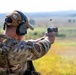 A military or law enforcement personnel is firing a semi-automatic pistol at an outdoor shooting range. A spent brass casing is visibly ejecting from the firearm, captured mid-air by the camera. The individual is wearing full tactical gear including a plate carrier, multicam uniform, hearing protection, and a baseball cap.