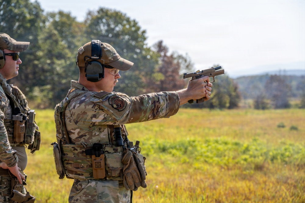 Two individuals in military/tactical camouflage uniforms are engaged in a firearms training exercise at an outdoor range. The primary subject is in a shooting stance, extending a tan/FDE-colored semi-automatic pistol with both hands. The setting appears to be a grassy field with trees in the background during daylight hours.