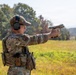 Two individuals in military/tactical camouflage uniforms are engaged in a firearms training exercise at an outdoor range. The primary subject is in a shooting stance, extending a tan/FDE-colored semi-automatic pistol with both hands. The setting appears to be a grassy field with trees in the background during daylight hours.