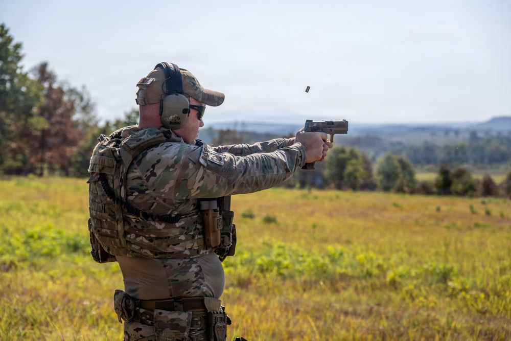 A person in full military/tactical camouflage gear is firing a semi-automatic pistol at an outdoor shooting range or training area. A spent brass casing is visibly ejecting from the firearm, captured mid-air. The setting appears to be an open field with trees and hills in the background under an overcast sky.