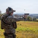 A person in full military/tactical camouflage gear is firing a semi-automatic pistol at an outdoor shooting range or training area. A spent brass casing is visibly ejecting from the firearm, captured mid-air. The setting appears to be an open field with trees and hills in the background under an overcast sky.