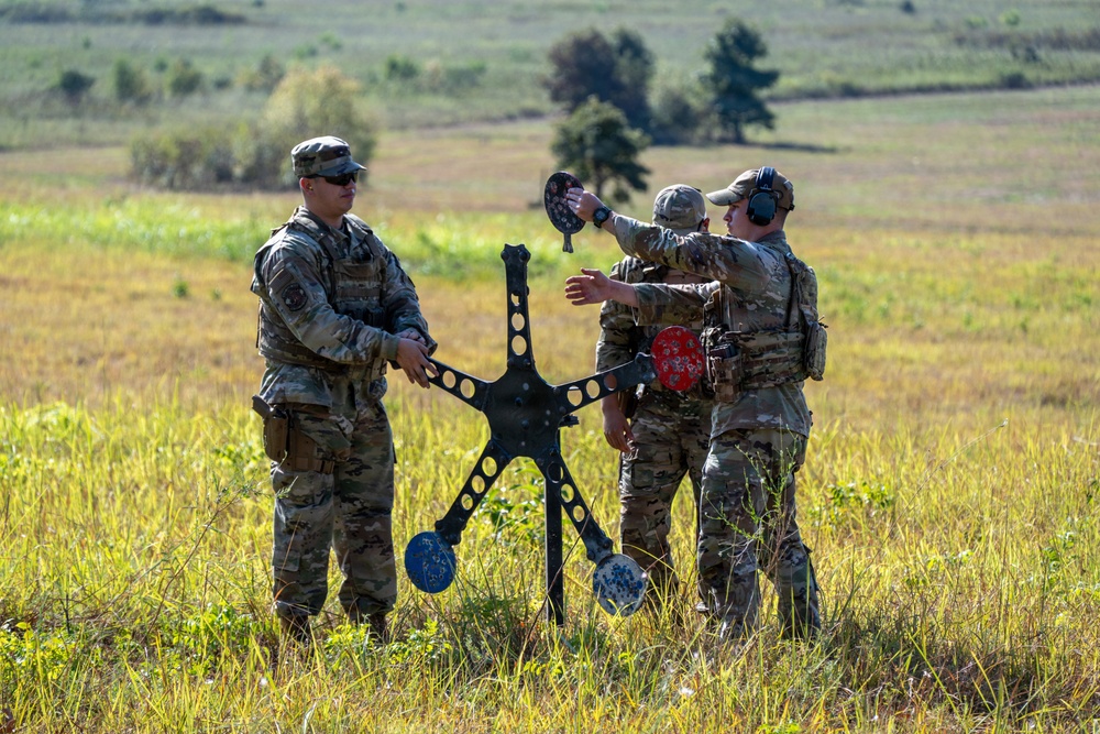 Three military personnel in camouflage uniforms are working with a metal shooting target stand in an open field. The target apparatus features multiple circular steel plate targets in blue, red, and black, showing evidence of bullet impacts. The soldiers appear to be setting up, adjusting, or resetting the target system during a live-fire training exercise.