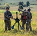 Three military personnel in camouflage uniforms are working with a metal shooting target stand in an open field. The target apparatus features multiple circular steel plate targets in blue, red, and black, showing evidence of bullet impacts. The soldiers appear to be setting up, adjusting, or resetting the target system during a live-fire training exercise.