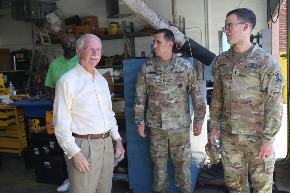 A civilian man in a light yellow dress shirt and khaki pants is engaged in a friendly conversation with two U.S. Army soldiers in OCP (Operational Camouflage Pattern) uniforms inside what appears to be a military maintenance or supply facility. A fourth individual in a green polo shirt is visible in the background. The setting suggests an official visit or inspection of a military workshop or storage area.