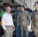A civilian man in a light yellow dress shirt and khaki pants is engaged in a friendly conversation with two U.S. Army soldiers in OCP (Operational Camouflage Pattern) uniforms inside what appears to be a military maintenance or supply facility. A fourth individual in a green polo shirt is visible in the background. The setting suggests an official visit or inspection of a military workshop or storage area.