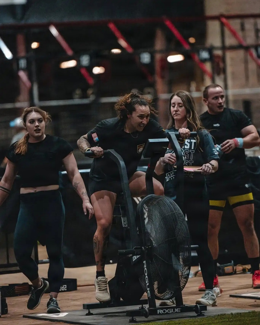 A fitness competition event featuring athletes performing on exercise equipment in an indoor arena setting. A central female athlete is actively using a Rogue Echo air bike while others stand nearby, appearing to be teammates or judges. The setting suggests a CrossFit-style competition with professional lighting and equipment.