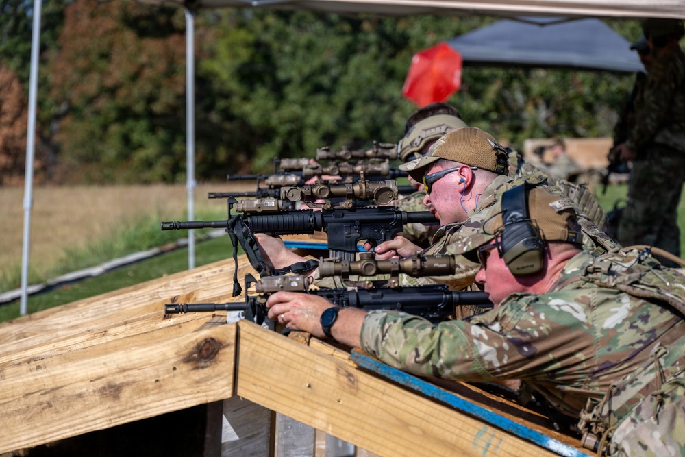 Multiple personnel in military/tactical camouflage uniforms are positioned at a wooden shooting bench, aiming rifles downrange. The setting appears to be an outdoor shooting range or training exercise, with canopy tents visible in the background. Shooters are equipped with optics-mounted rifles, hearing protection, and tactical gear.