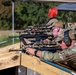Multiple personnel in military/tactical camouflage uniforms are positioned at a wooden shooting bench, aiming rifles downrange. The setting appears to be an outdoor shooting range or training exercise, with canopy tents visible in the background. Shooters are equipped with optics-mounted rifles, hearing protection, and tactical gear.