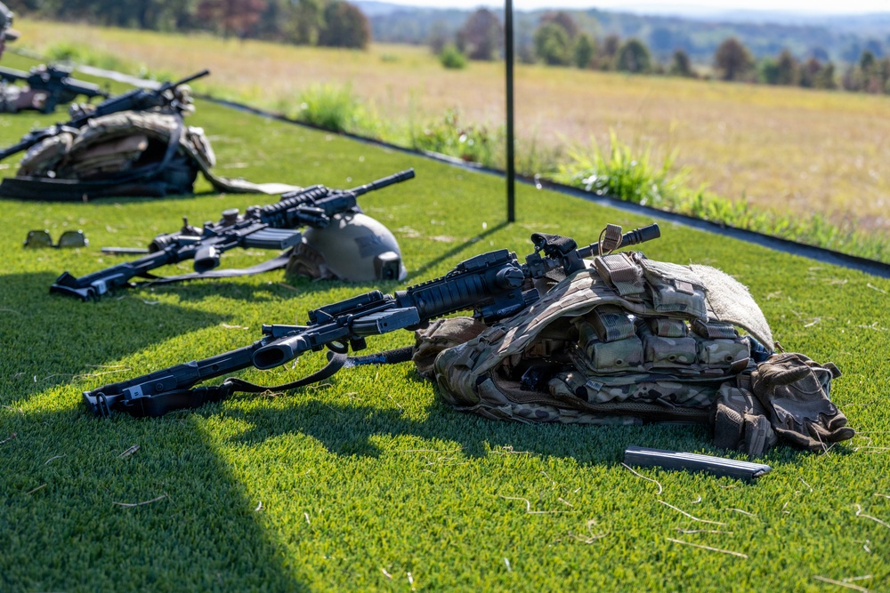 Military equipment is laid out on a grass surface, likely at a training range or equipment display area. Multiple firearms, tactical vests, helmets, and gear are arranged on the ground in an organized manner. The setting appears to be an outdoor military training or demonstration event.