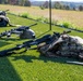 Military equipment is laid out on a grass surface, likely at a training range or equipment display area. Multiple firearms, tactical vests, helmets, and gear are arranged on the ground in an organized manner. The setting appears to be an outdoor military training or demonstration event.