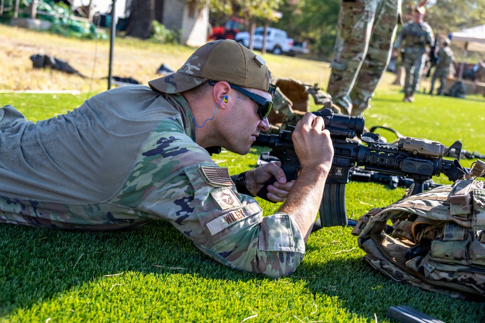 A U.S. Air Force service member identified by name tape as 'WILEY' is in a prone shooting position on grass, aiming through the optic of an M4-style carbine rifle. The individual is wearing OCP (Operational Camouflage Pattern) uniform, a tan ball cap, sunglasses, and foam ear protection. Additional military personnel in OCP uniforms are visible standing in the background, suggesting a training or qualification range event.
