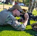 A U.S. Air Force service member identified by name tape as 'WILEY' is in a prone shooting position on grass, aiming through the optic of an M4-style carbine rifle. The individual is wearing OCP (Operational Camouflage Pattern) uniform, a tan ball cap, sunglasses, and foam ear protection. Additional military personnel in OCP uniforms are visible standing in the background, suggesting a training or qualification range event.