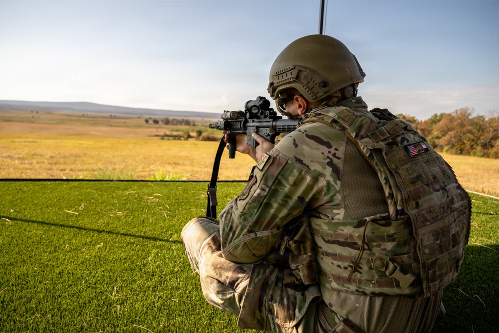 A military soldier in full combat gear is kneeling in a firing position, aiming a rifle with an optical sight across an open field. The soldier is wearing MultiCam camouflage, a tactical helmet, body armor/plate carrier, and has visible flag patches on the right shoulder. The setting appears to be a training range or observation post with a grassy foreground and open terrain in the background.