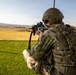 A military soldier in full combat gear is kneeling in a firing position, aiming a rifle with an optical sight across an open field. The soldier is wearing MultiCam camouflage, a tactical helmet, body armor/plate carrier, and has visible flag patches on the right shoulder. The setting appears to be a training range or observation post with a grassy foreground and open terrain in the background.