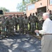 A civilian man in a white long-sleeve shirt and khaki pants addresses a large group of military personnel in camouflage uniforms outside a brick building. The soldiers are standing in an informal assembly on a gravel/asphalt surface, listening to the speaker who is holding a water bottle. The setting appears to be a military installation or armory during daytime.