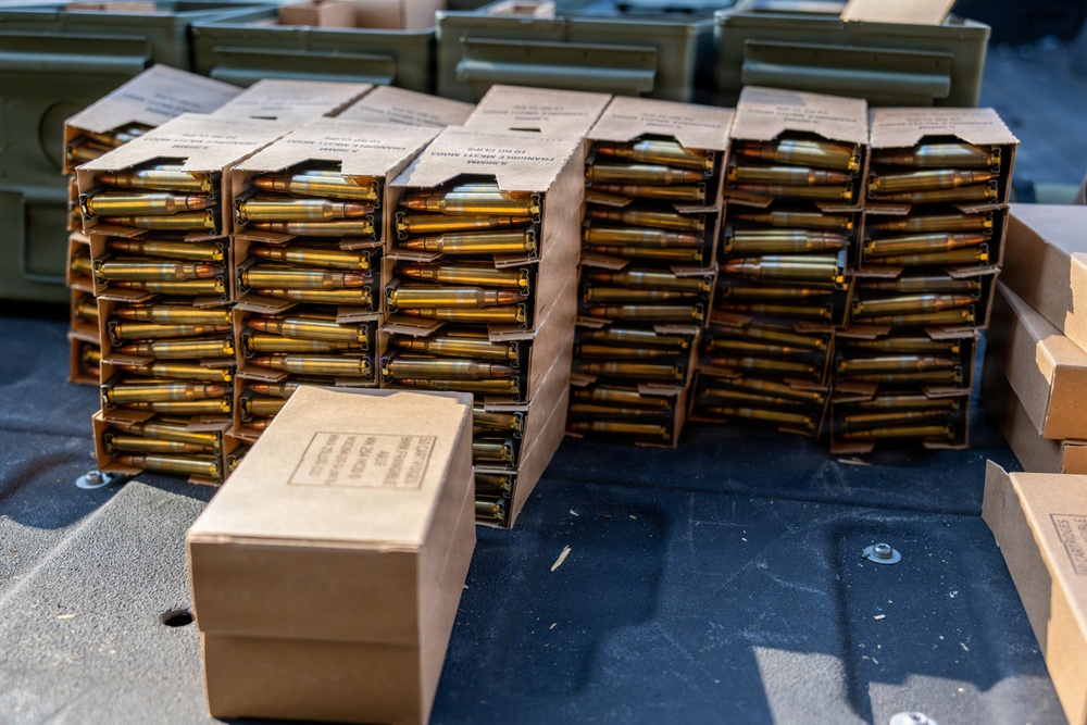 The image shows a large quantity of rifle ammunition organized in cardboard stripper clip packaging, stacked in multiple columns on a dark surface. The cartridges appear to be military-style rifle rounds with brass casings and copper-jacketed projectiles. Green military ammunition cans are visible in the background.