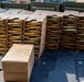 The image shows a large quantity of rifle ammunition organized in cardboard stripper clip packaging, stacked in multiple columns on a dark surface. The cartridges appear to be military-style rifle rounds with brass casings and copper-jacketed projectiles. Green military ammunition cans are visible in the background.