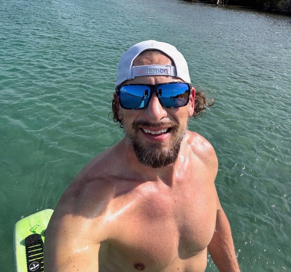 A shirtless man is taking a selfie near a body of calm, turquoise-green water. He is smiling broadly and wearing blue mirrored sunglasses and a white Lululemon snapback cap worn backwards. A green board, likely a wakeboard or bodyboard, is partially visible in the lower left corner.