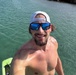 A shirtless man is taking a selfie near a body of calm, turquoise-green water. He is smiling broadly and wearing blue mirrored sunglasses and a white Lululemon snapback cap worn backwards. A green board, likely a wakeboard or bodyboard, is partially visible in the lower left corner.