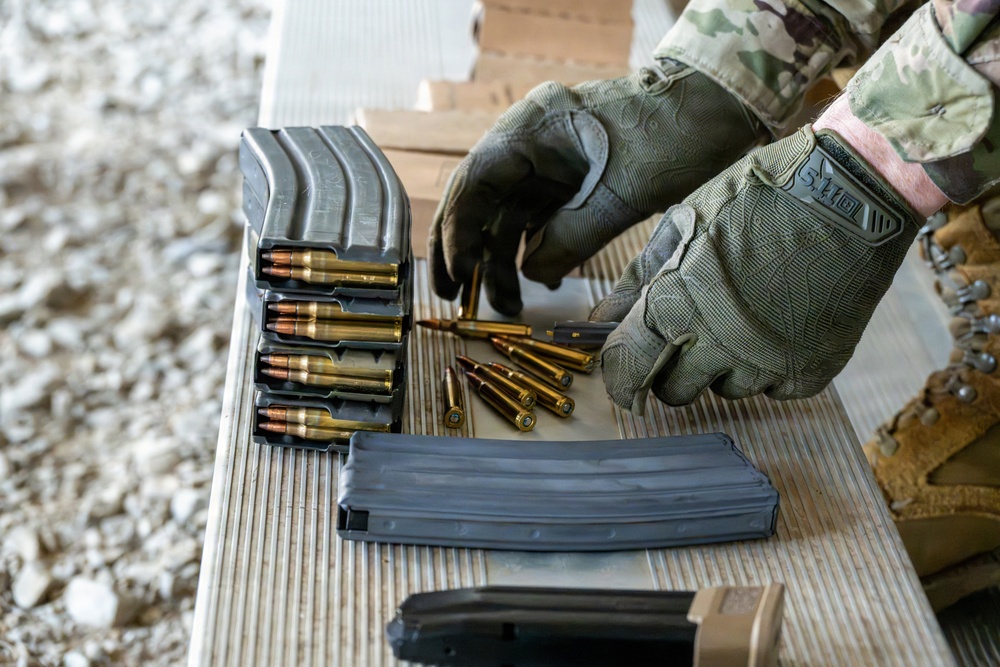 A person wearing military multicam uniform and 5.11 tactical gloves is loading ammunition magazines on a wooden surface at what appears to be a shooting range. Multiple AR-style rifle magazines are visible, some already loaded with rifle cartridges, while loose rounds are scattered on the surface. The activity suggests a pre-range preparation or magazine loading drill.