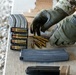A person wearing military multicam uniform and 5.11 tactical gloves is loading ammunition magazines on a wooden surface at what appears to be a shooting range. Multiple AR-style rifle magazines are visible, some already loaded with rifle cartridges, while loose rounds are scattered on the surface. The activity suggests a pre-range preparation or magazine loading drill.