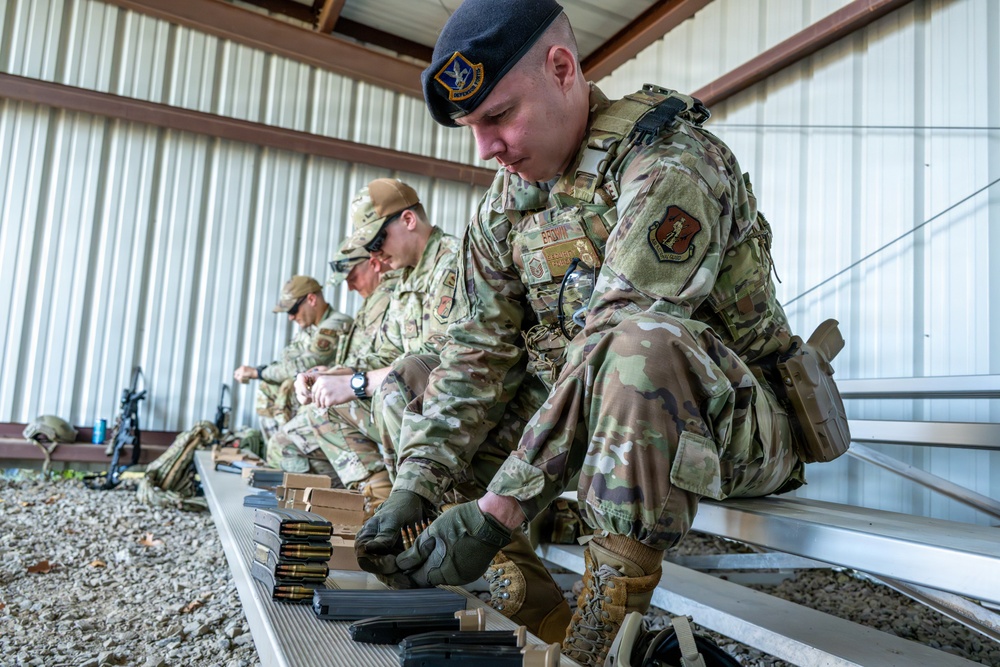 Multiple military personnel in OCP (Operational Camouflage Pattern) uniforms are seated on a metal bench under a covered range shelter, loading ammunition magazines. The foreground soldier wears a black beret with a unit flash and tactical gear including a holster and plate carrier. The setting appears to be a military shooting range during a training exercise.