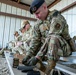 Multiple military personnel in OCP (Operational Camouflage Pattern) uniforms are seated on a metal bench under a covered range shelter, loading ammunition magazines. The foreground soldier wears a black beret with a unit flash and tactical gear including a holster and plate carrier. The setting appears to be a military shooting range during a training exercise.