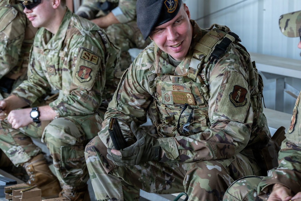 The image shows multiple U.S. Air Force Security Forces personnel in OCP (Operational Camouflage Pattern) uniforms seated together, likely during a briefing or rest period. The central figure, identified by a name tape reading 'BROWN,' is wearing a beret with a unit insignia, a tactical vest, and tactical gloves, and appears to be smiling. The personnel display Security Forces ('SF') patches and unit insignia patches on their uniforms.