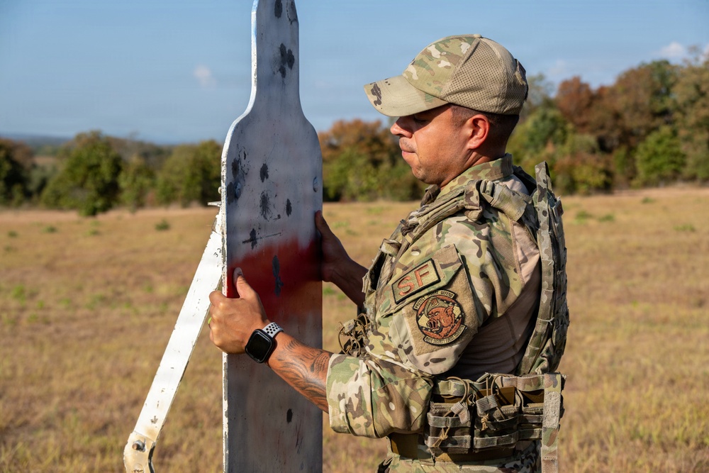 A military or security forces personnel wearing OCP (Operational Camouflage Pattern) uniform and tactical vest is examining a bullet-riddled shooting range target. The individual displays 'SF' (Security Forces) patches on their uniform, indicating US Air Force Security Forces affiliation. The scene appears to be an outdoor firing range with an open field and tree line in the background.