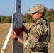 A military or security forces personnel wearing OCP (Operational Camouflage Pattern) uniform and tactical vest is examining a bullet-riddled shooting range target. The individual displays 'SF' (Security Forces) patches on their uniform, indicating US Air Force Security Forces affiliation. The scene appears to be an outdoor firing range with an open field and tree line in the background.