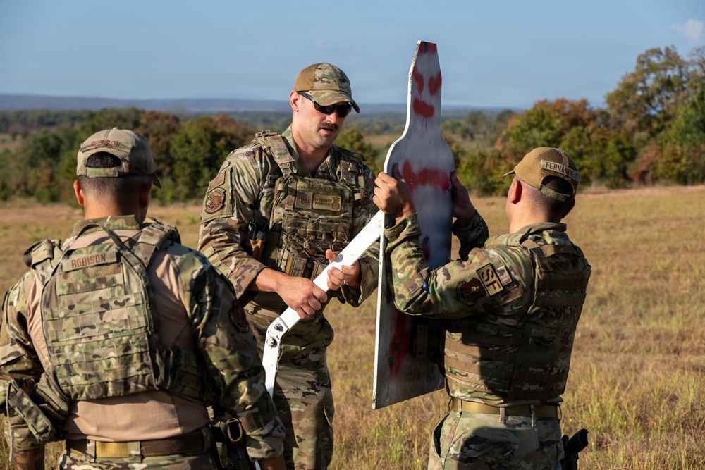 Three military personnel in OCP (Operational Camouflage Pattern) uniforms are gathered in an open field examining a white shooting target with red paint markings indicating bullet impacts. The central figure, wearing sunglasses and a tactical vest, appears to be an instructor reviewing target results with trainees. The setting appears to be a military training range during daylight hours.