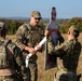 Three military personnel in OCP (Operational Camouflage Pattern) uniforms are gathered in an open field examining a white shooting target with red paint markings indicating bullet impacts. The central figure, wearing sunglasses and a tactical vest, appears to be an instructor reviewing target results with trainees. The setting appears to be a military training range during daylight hours.