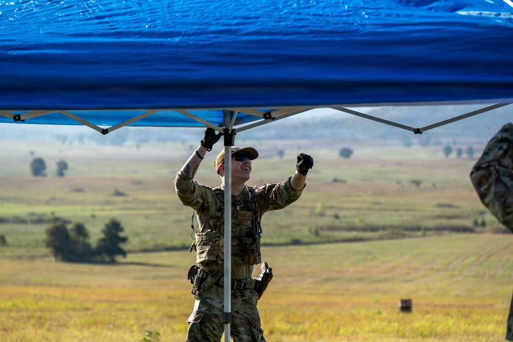 A military service member in camouflage uniform and tactical gear is setting up or adjusting a blue pop-up canopy tent in an open field environment. The individual is wearing black gloves and a tan cap while looking upward at the tent frame. The background reveals a wide, flat grassy landscape consistent with a training range or field exercise area.