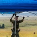 A military service member in camouflage uniform and tactical gear is setting up or adjusting a blue pop-up canopy tent in an open field environment. The individual is wearing black gloves and a tan cap while looking upward at the tent frame. The background reveals a wide, flat grassy landscape consistent with a training range or field exercise area.