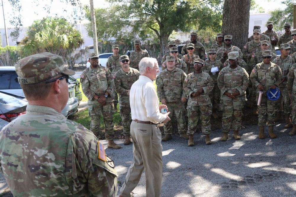 A civilian in a white dress shirt and khaki pants addresses a large group of U.S. Army soldiers in OCP (Operational Camouflage Pattern) uniforms outdoors. The gathering appears to be an informal briefing or address, possibly at a military installation or staging area. A soldier with a name tape reading 'KLEIN' is visible in the foreground on the left.