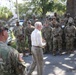A civilian in a white dress shirt and khaki pants addresses a large group of U.S. Army soldiers in OCP (Operational Camouflage Pattern) uniforms outdoors. The gathering appears to be an informal briefing or address, possibly at a military installation or staging area. A soldier with a name tape reading 'KLEIN' is visible in the foreground on the left.