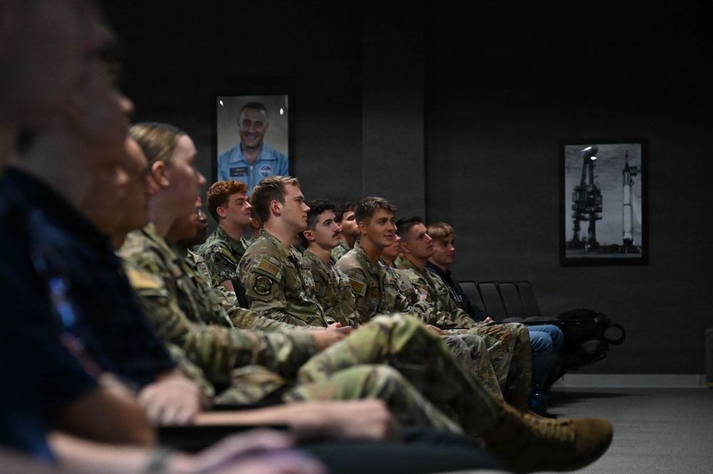 A group of military personnel in camouflage uniforms are seated in rows, appearing to attend a briefing or presentation in a formal room. The setting features framed photographs on the wall, including what appears to be a NASA astronaut portrait and a black-and-white rocket launch pad image. The audience appears attentive and engaged, facing toward an unseen presenter or screen.