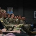 A group of military personnel in camouflage uniforms are seated in rows, appearing to attend a briefing or presentation in a formal room. The setting features framed photographs on the wall, including what appears to be a NASA astronaut portrait and a black-and-white rocket launch pad image. The audience appears attentive and engaged, facing toward an unseen presenter or screen.