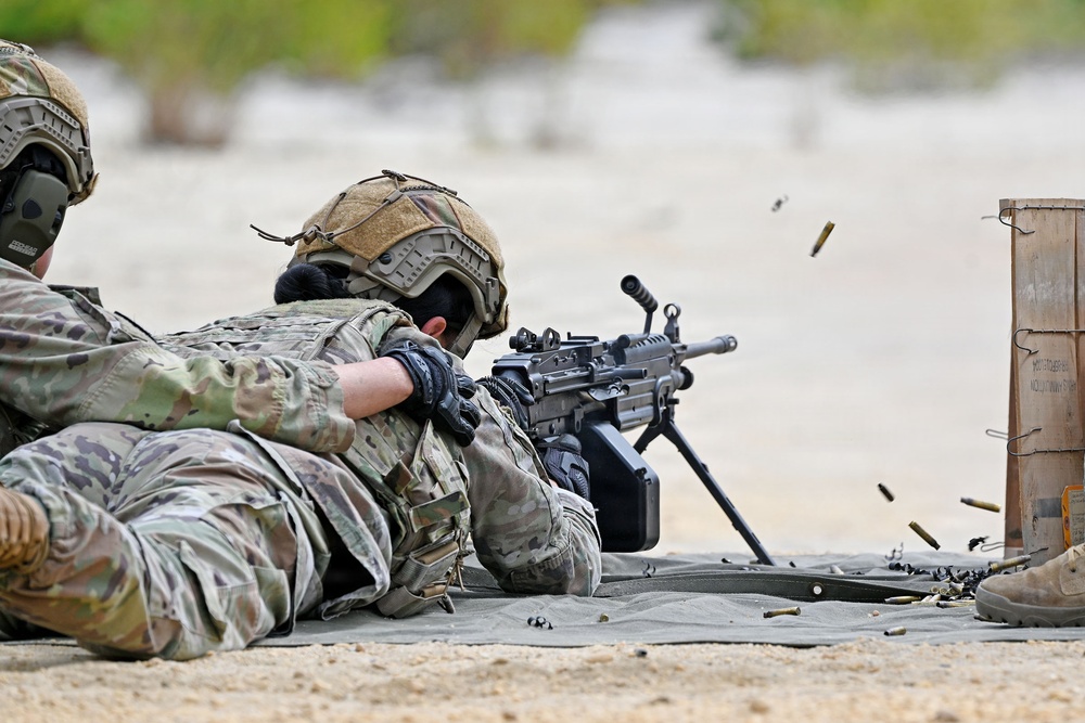 Two military personnel in full combat gear are conducting a live-fire training exercise in a prone position on a sandy range. The shooter in the foreground is actively firing a light machine gun, with spent brass casings visibly ejecting from the weapon. A wooden ammunition crate is positioned to the right side of the firing position.